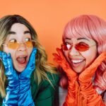 Two joyful women in colorful wigs and gloves expressing surprise on an orange background.